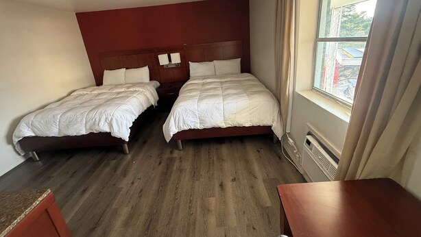 A hotel room with two beds, a wooden floor, a window with curtains, and a small table.