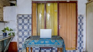 Una habitación con una mesa cubierta por un mantel estampado, una laptop sobre la mesa, una planta en maceta y una puerta con cortina amarilla.