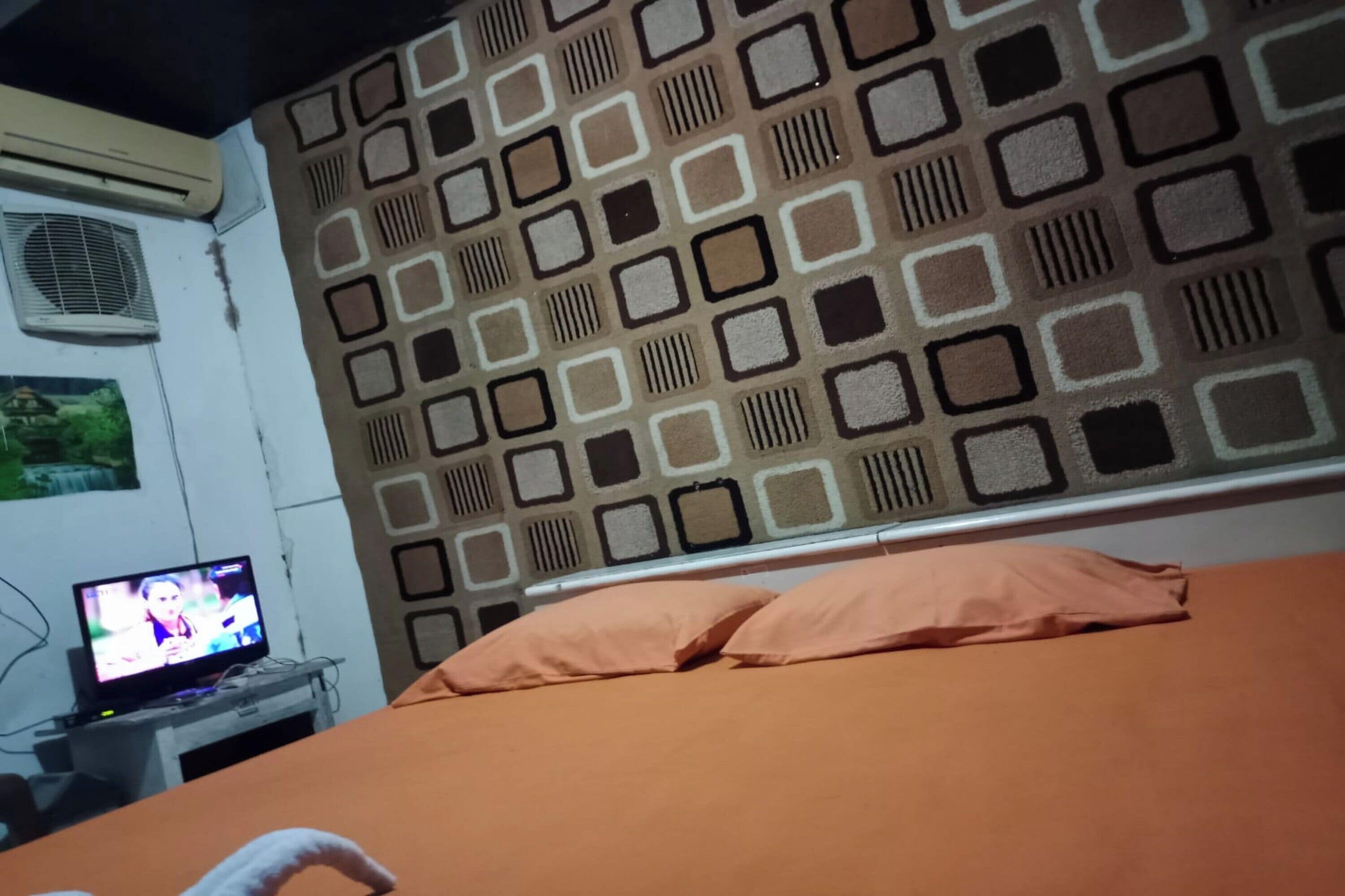A room with a patterned wall, a bed with orange bedding, a television on a stand, and an air conditioning unit.