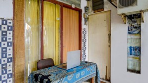 Una habitación con una laptop sobre una mesa, una silla, un mantel estampado, piso de baldosas y una puerta con un calendario en la pared.