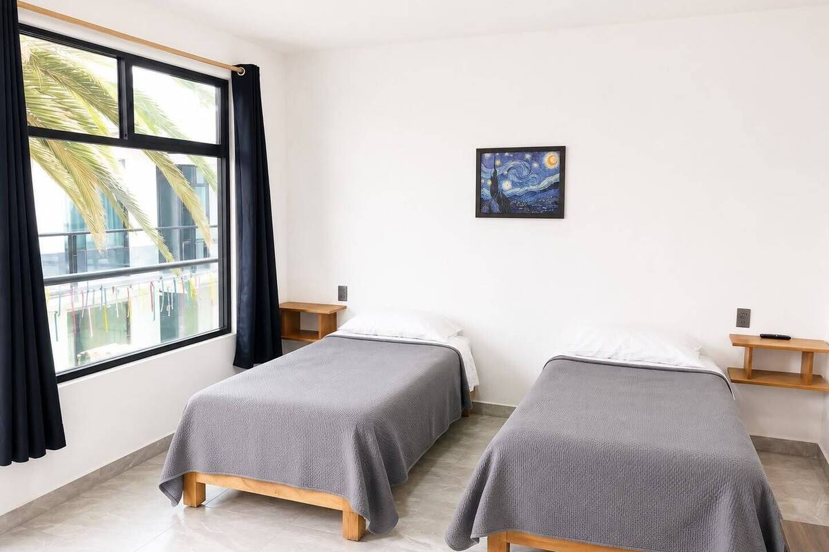 A room with two beds, a window with curtains, a painting on the wall, and a small wooden nightstand.