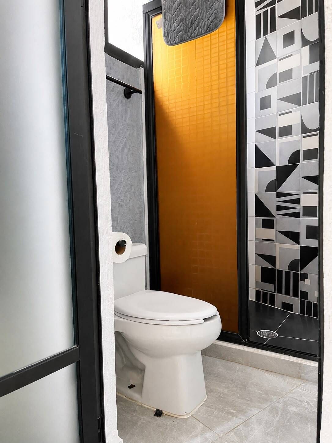 Um banheiro com vaso sanitário branco, porta-toalhas e área de chuveiro com porta de padrão geométrico.