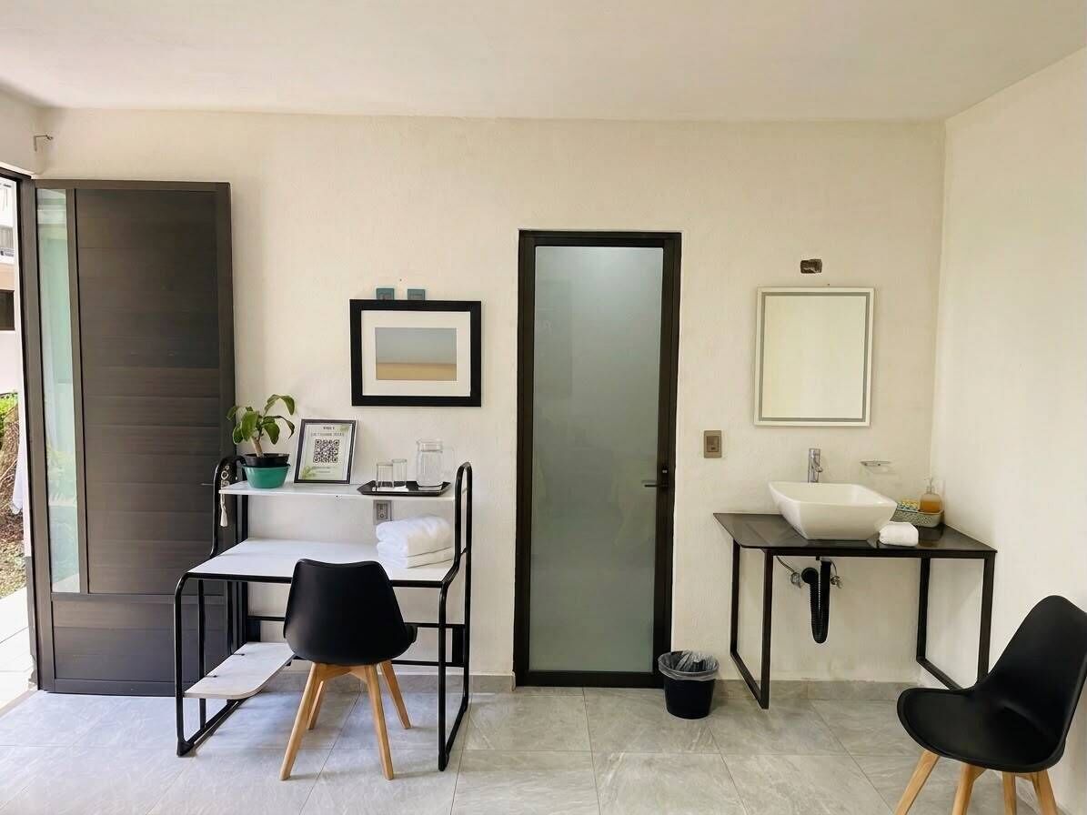 Una habitación pequeña y moderna con un escritorio blanco, dos sillas negras, un lavamanos blanco, un espejo y una puerta.