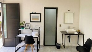 Une petite pièce moderne avec un bureau blanc, deux chaises noires, un lavabo blanc, un miroir et une porte.