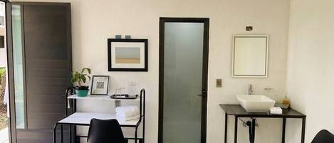 Une petite pièce moderne avec un bureau blanc, deux chaises noires, un lavabo blanc, un miroir et une porte.