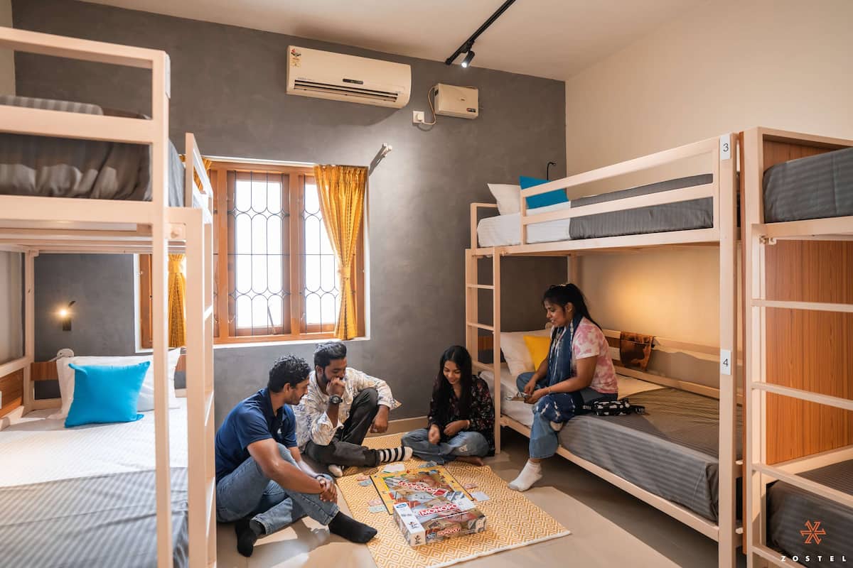 A bunk bed room with a window, air conditioning unit, and a group of people sitting on the floor playing a board game.