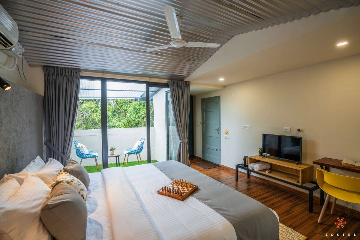 A modern hotel room with a large bed, a TV, a desk, and a balcony with chairs.
