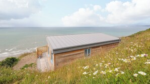 Un edificio costiero con tetto in metallo, pareti in legno e un piccolo portico. Si affaccia sul mare ed è circondato da fiori selvatici.