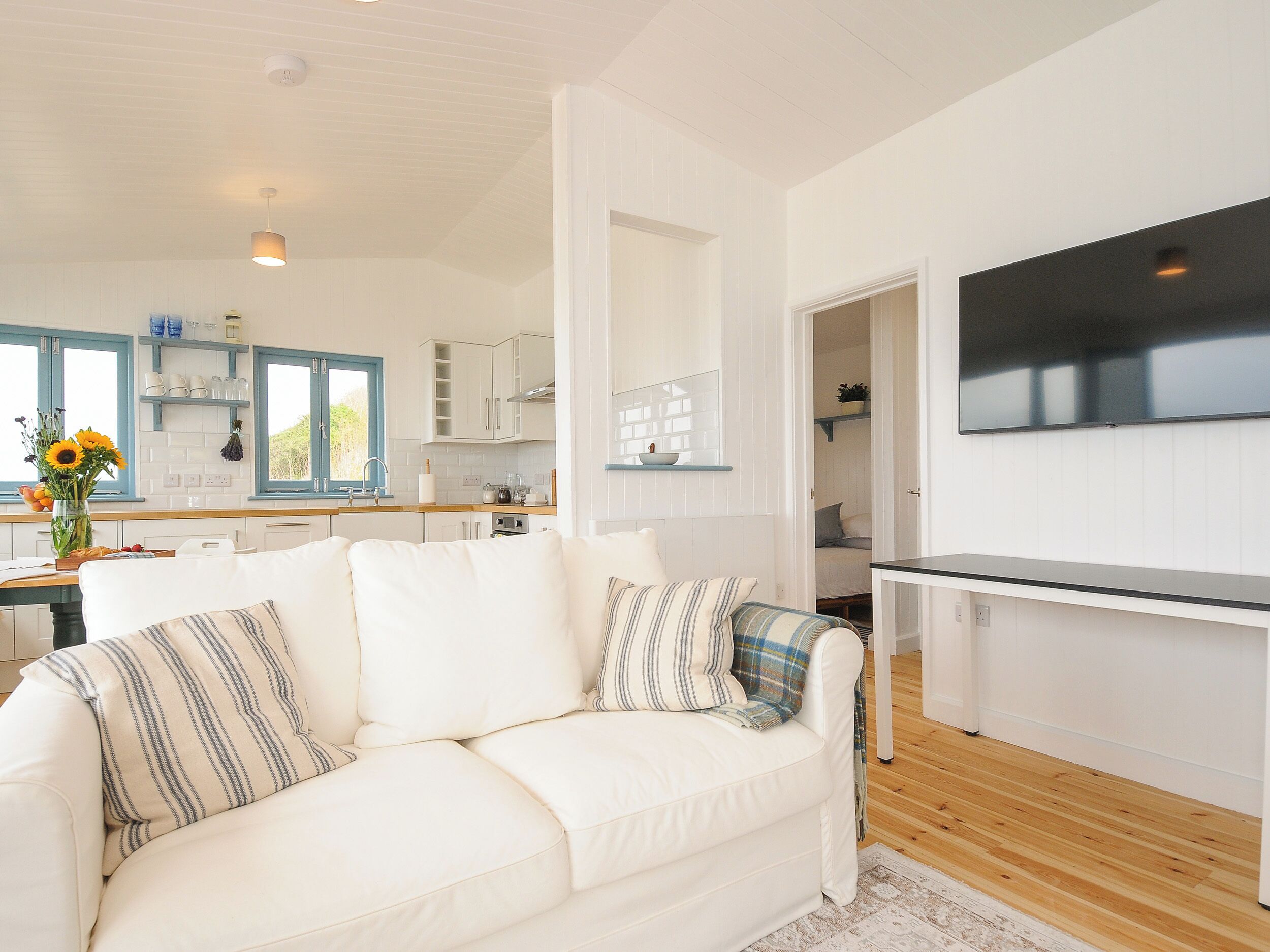 A modern, well-lit living room with a white sofa, a flat-screen TV, and a kitchen in the background.