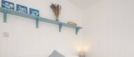 A neatly made bed with white bedding and a single blue pillow, a small bedside table with a lamp, and a wall-mounted shelf with decorative items.
