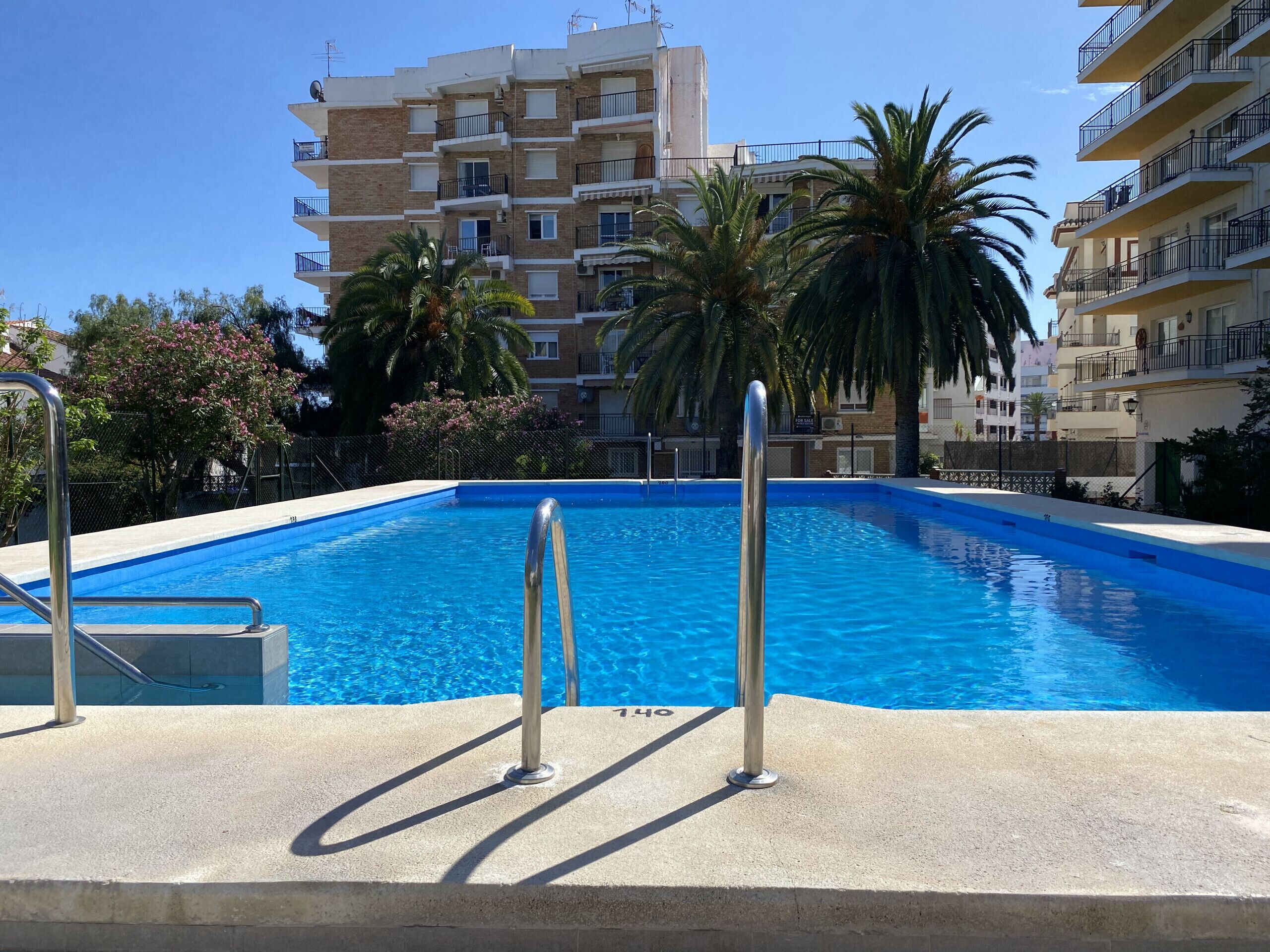 A swimming pool with metal railings, surrounded by a concrete deck. In the background, there are multi-story buildings with balconies and palm trees.