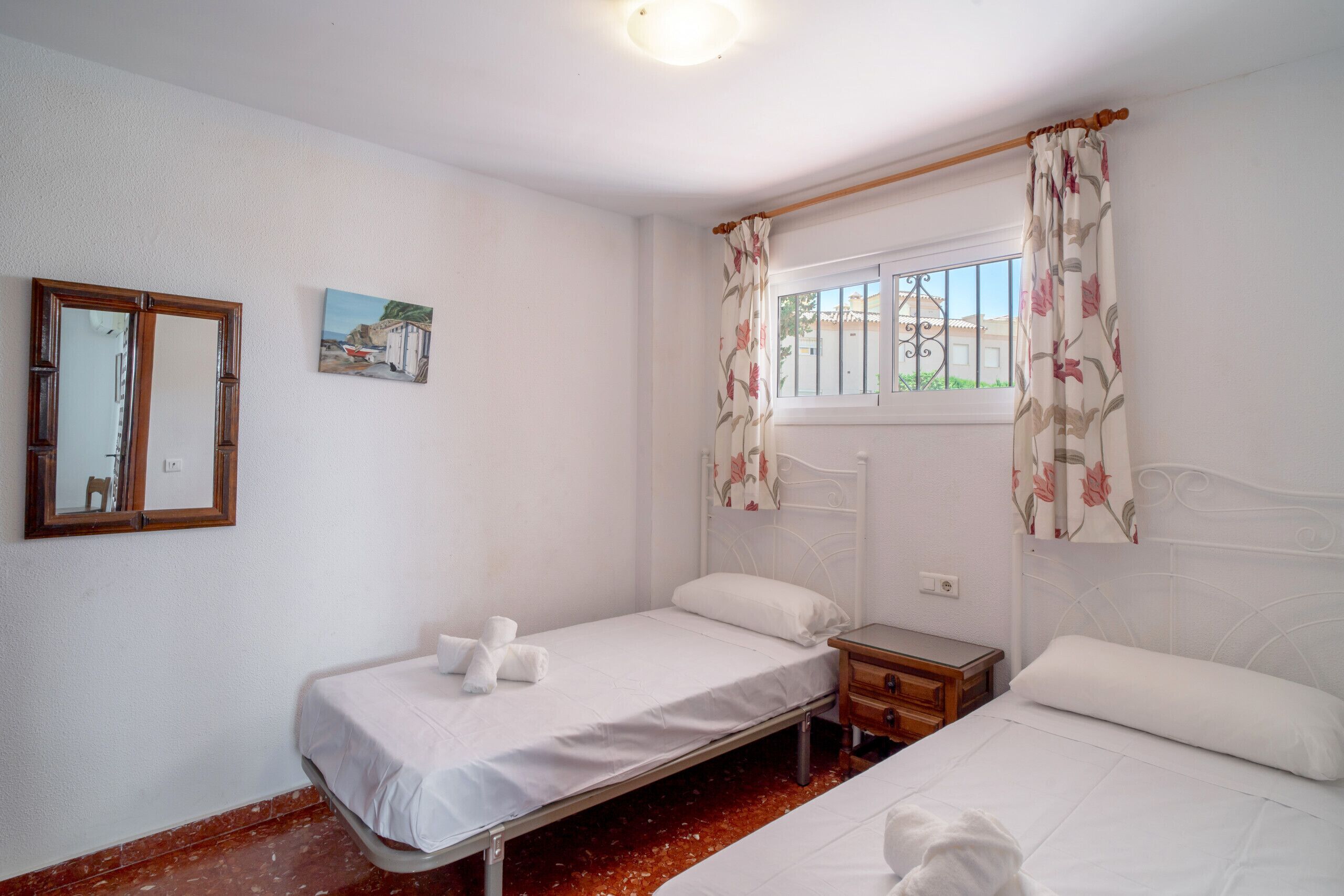 A room with two beds, a wooden nightstand, a mirror, and a window with floral curtains.