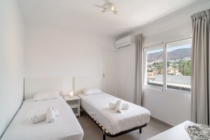 A hotel room with two single beds, a bedside table with a lamp, an air conditioning unit, and a window with a view of a cityscape. - Perdigon 325 Paradise by Casasol (Nerja)