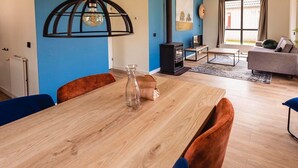 A modern living room with a wooden dining table, blue chairs, a sofa, a fireplace, and a large window.