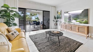 Uma sala de estar com sofá, mesa de centro, televisão e porta de vidro deslizante que dá para uma área externa com mesa e cadeiras.