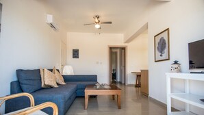 A living room with a blue sofa, a wooden coffee table, a ceiling fan, and a white TV stand. - Villa Eleftheria: 4 bedrooms for up to 9 adults, pool and outstanding 180° views (Lakithra)