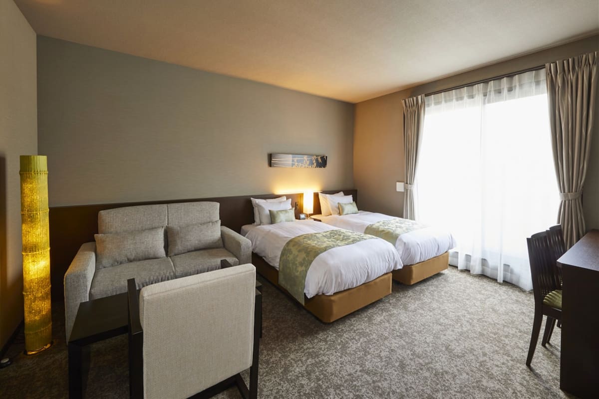 Habitación de hotel con sofá, dos camas, escritorio y un ventanal grande con cortinas.