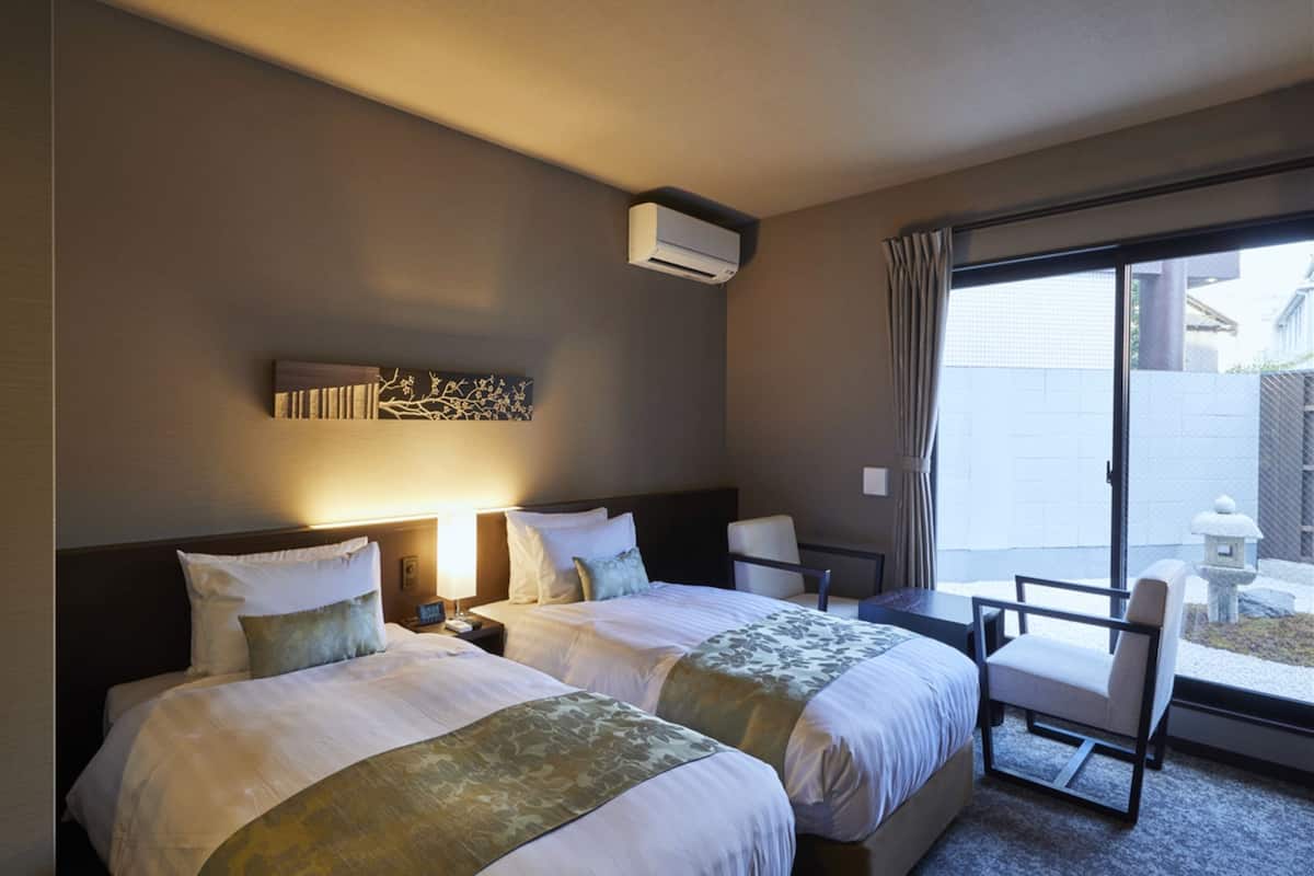 Habitación de hotel con dos camas, una mesita y una silla. Se aprecia una ventana con cortinas y vistas al exterior.