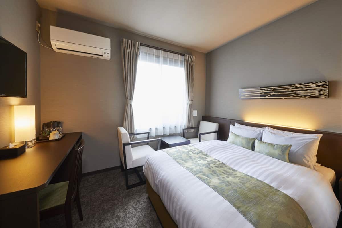 Una habitación de hotel con cama, escritorio, silla, televisor y ventana con cortinas.