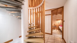 Una escalera de caracol con barandillas de madera y peldaños color beige que conduce a un dormitorio con cama y sillón.