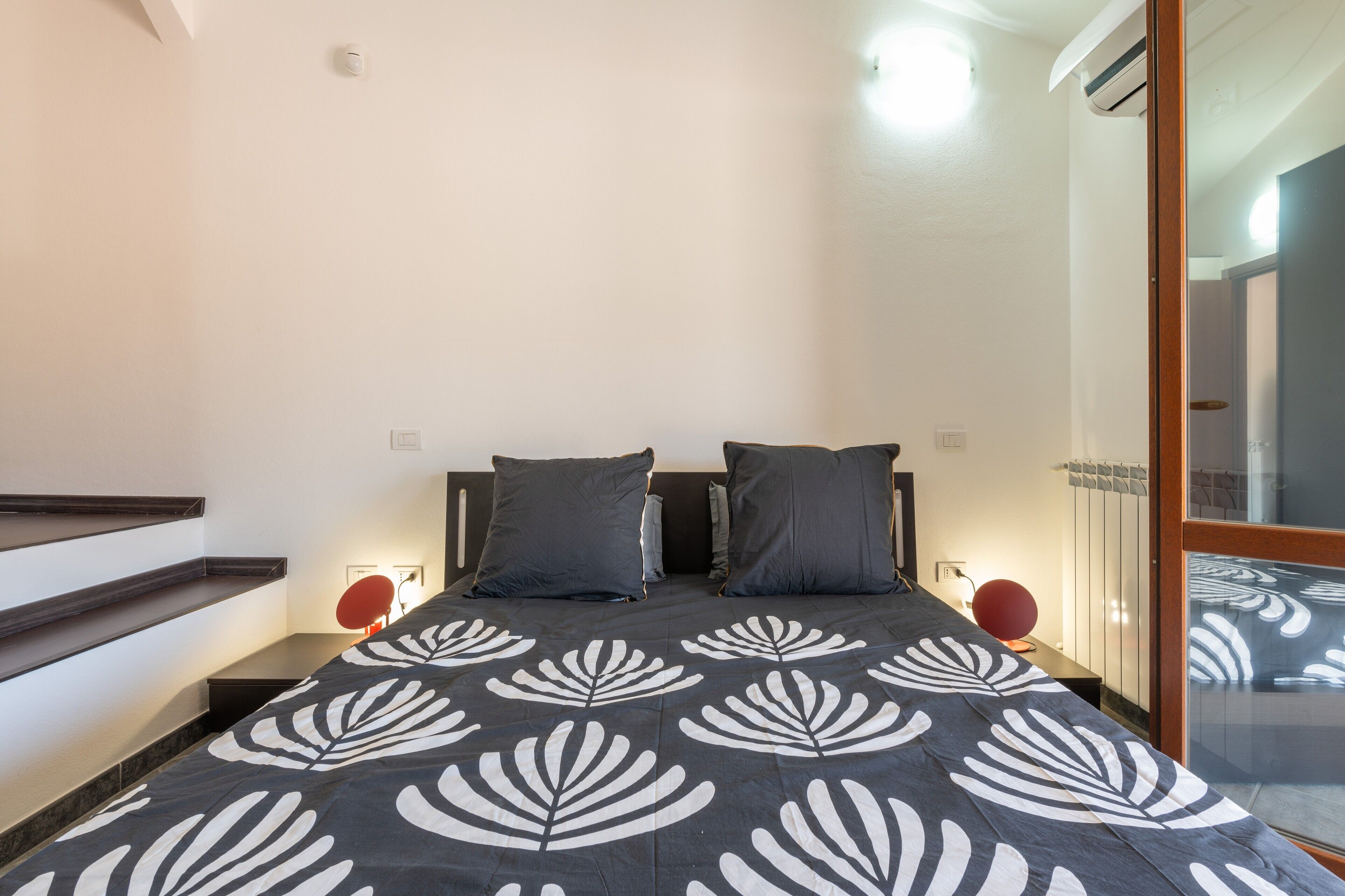 A bedroom with a bed, two pillows, a nightstand with a lamp, a mirror, and a sliding door.