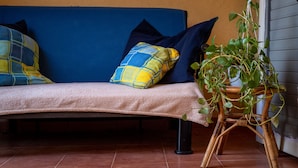 Een blauwe bank met een beige plaid, een geel-blauw kussen en een potplant op een bamboestandaard.