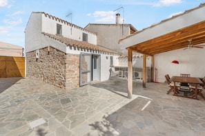 A stone patio leading to a white building with a tiled roof, a wooden pergola, and a dining area with chairs and a table.