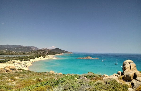 A coastal landscape with a beach, clear turquoise waters, and rocky outcrops.