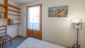 A bedroom with a bed, a chair, a wooden shelf unit, a window with a curtain, a wall-mounted lamp, and a painting.