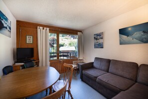 A room with a wooden dining table, a brown sofa, a television, and a window with curtains.