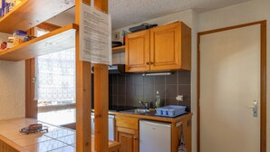 A compact kitchen with wooden cabinets, a small refrigerator, and a window with patterned curtains.