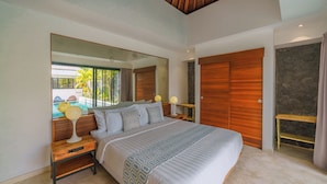 A bedroom with a large bed, a wooden headboard, a nightstand with a lamp, a large mirror, and a view of a swimming pool. - Villa Yasmee (Seminyak)
