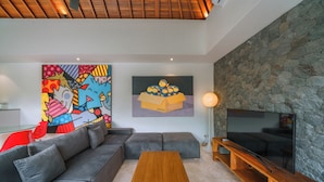 A modern living room with a grey sectional sofa, a wooden coffee table, a flat-screen TV on a wooden stand, a stone accent wall, and colorful abstract art on the walls. - Villa Yasmee (Seminyak)