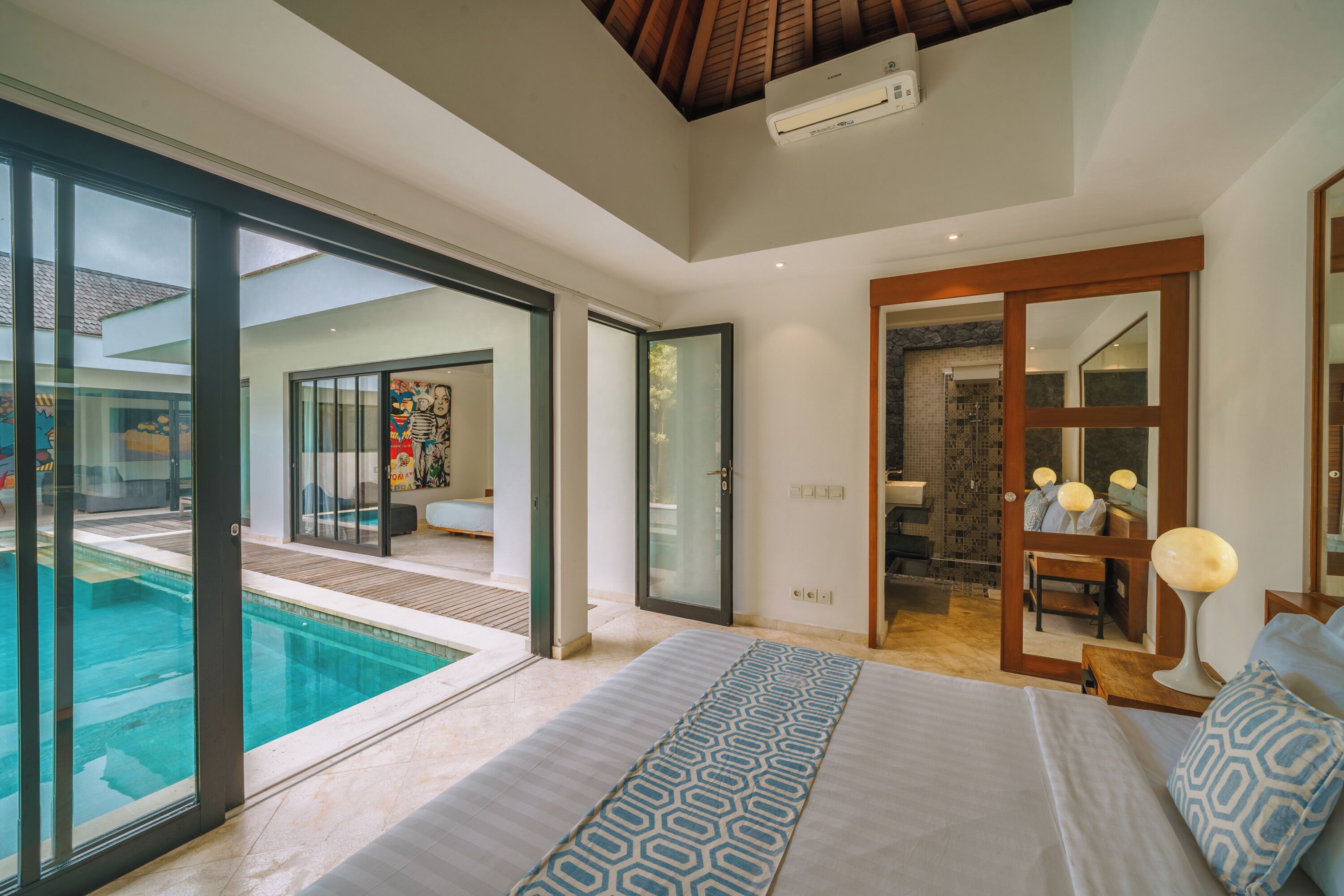 A modern hotel room with a large bed, a view of a swimming pool, and a bathroom with a shower and mirror.