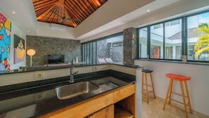 A modern kitchen with a black countertop, stainless steel sink, wooden cabinets, and a stone backsplash. There are two wooden stools and a large window providing a view of the outside. - Villa Yasmee (Seminyak)