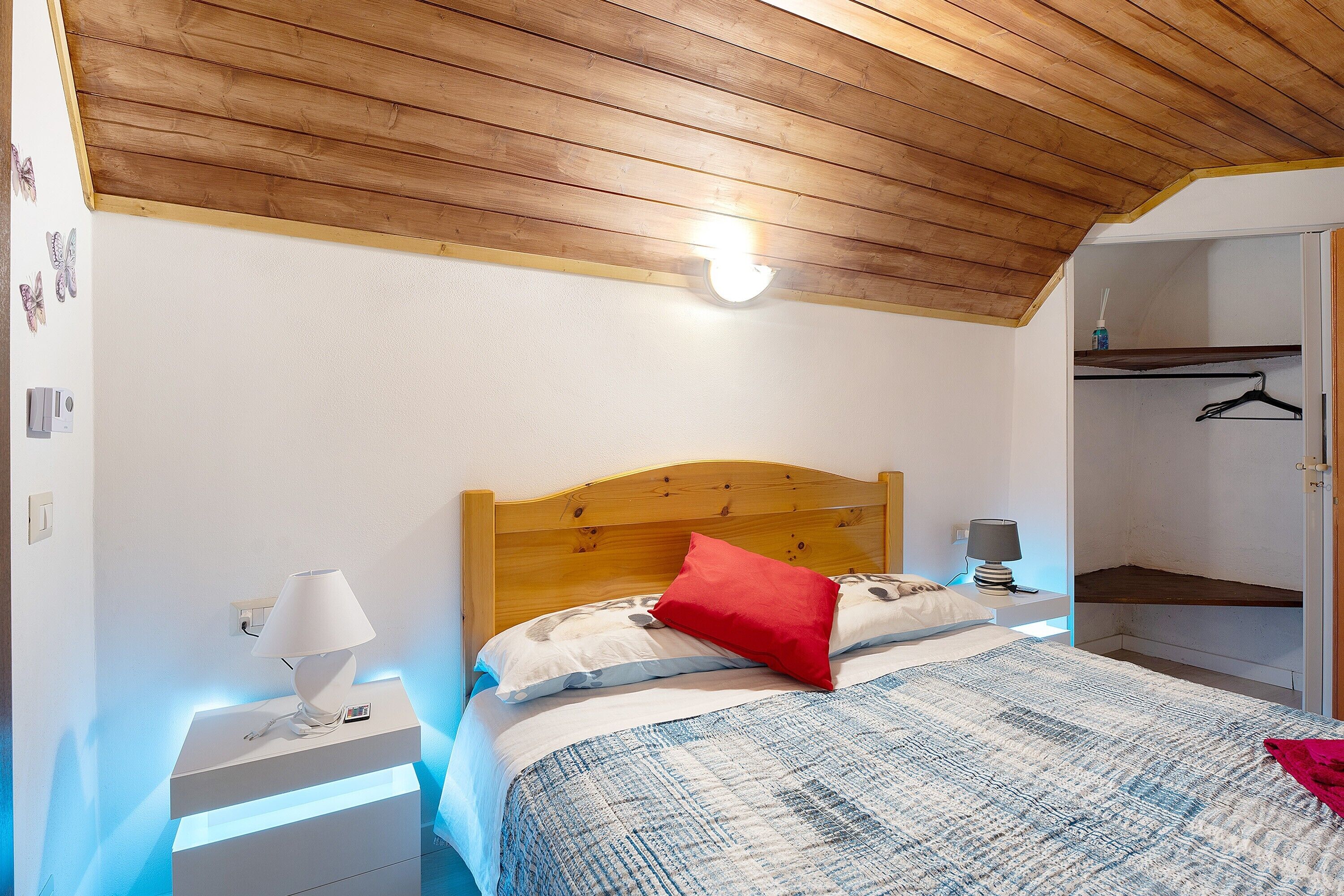 A bedroom with a wooden ceiling, a bed with a white headboard, a bedside table with a lamp, and a closet with a hanging rod.