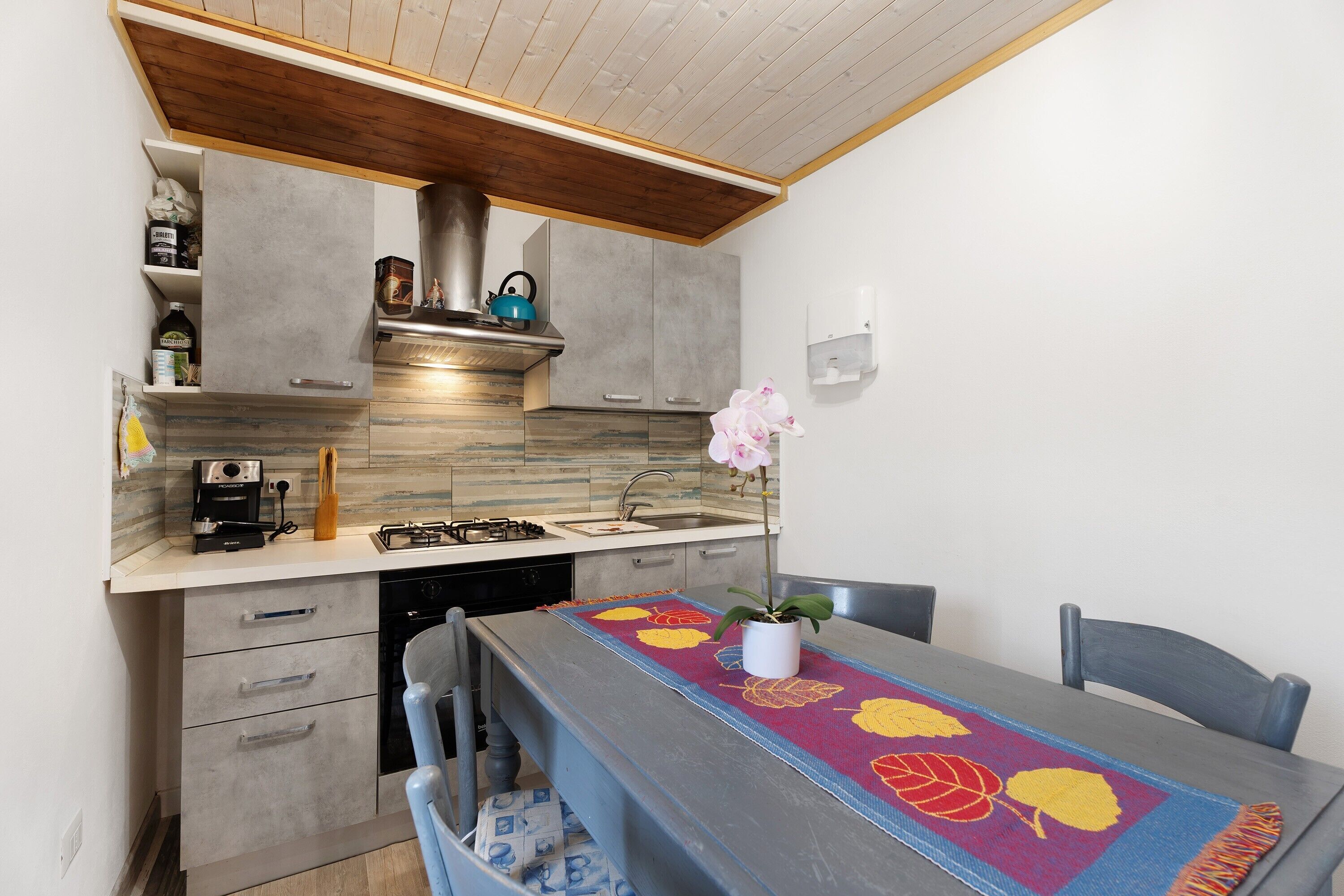 Une cuisine avec des meubles gris, une hotte en acier inoxydable, une bouilloire bleue et une table agrémentée d’un chemin de table à motifs floraux.