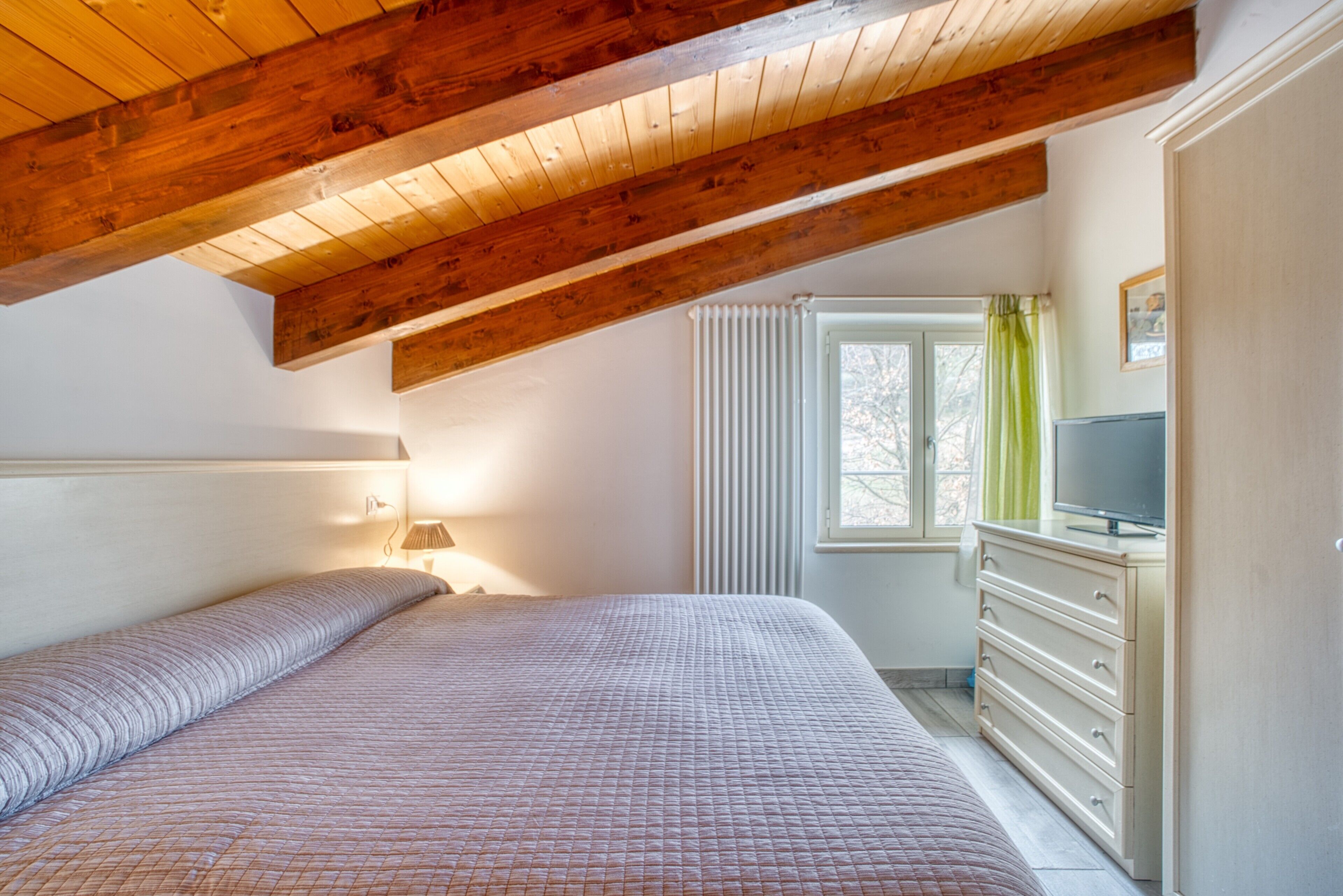 A bedroom with a wooden ceiling, a bed, a dresser, a television, and a window with curtains.