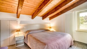 Ein Schlafzimmer mit Holzbalkendecke, einem Bett mit gemustertem Bettbezug, Nachttischlampen, einem Nachttisch und einem Fenster mit Jalousien.