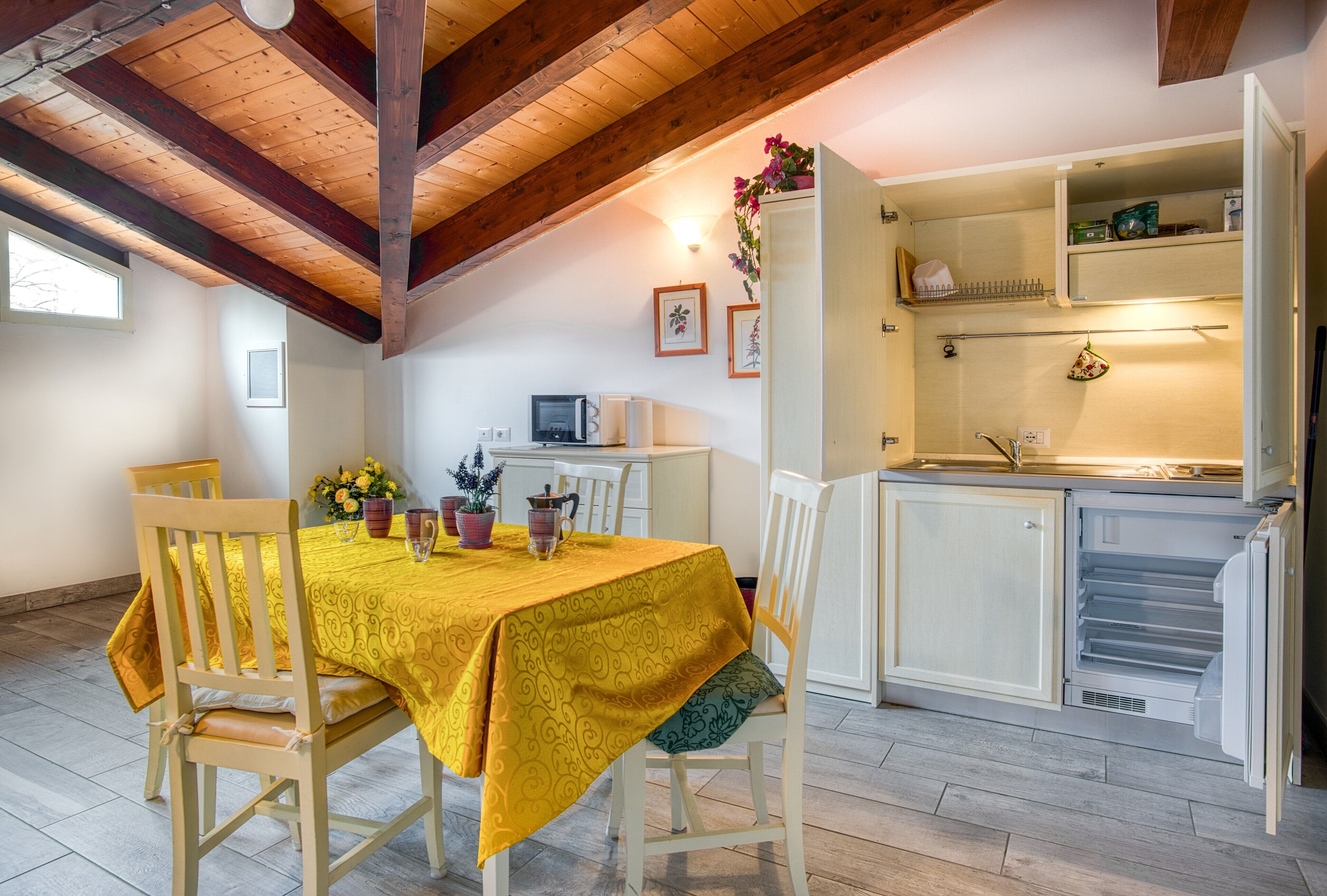 Een eetgedeelte met een gele tafelkleed, houten stoelen, een keuken met een witte koelkast en een houten plafond.