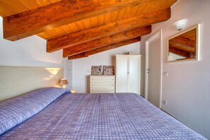 A bedroom with a wooden ceiling, a bed with a patterned bedspread, a dresser, a mirror, and wall-mounted lights.