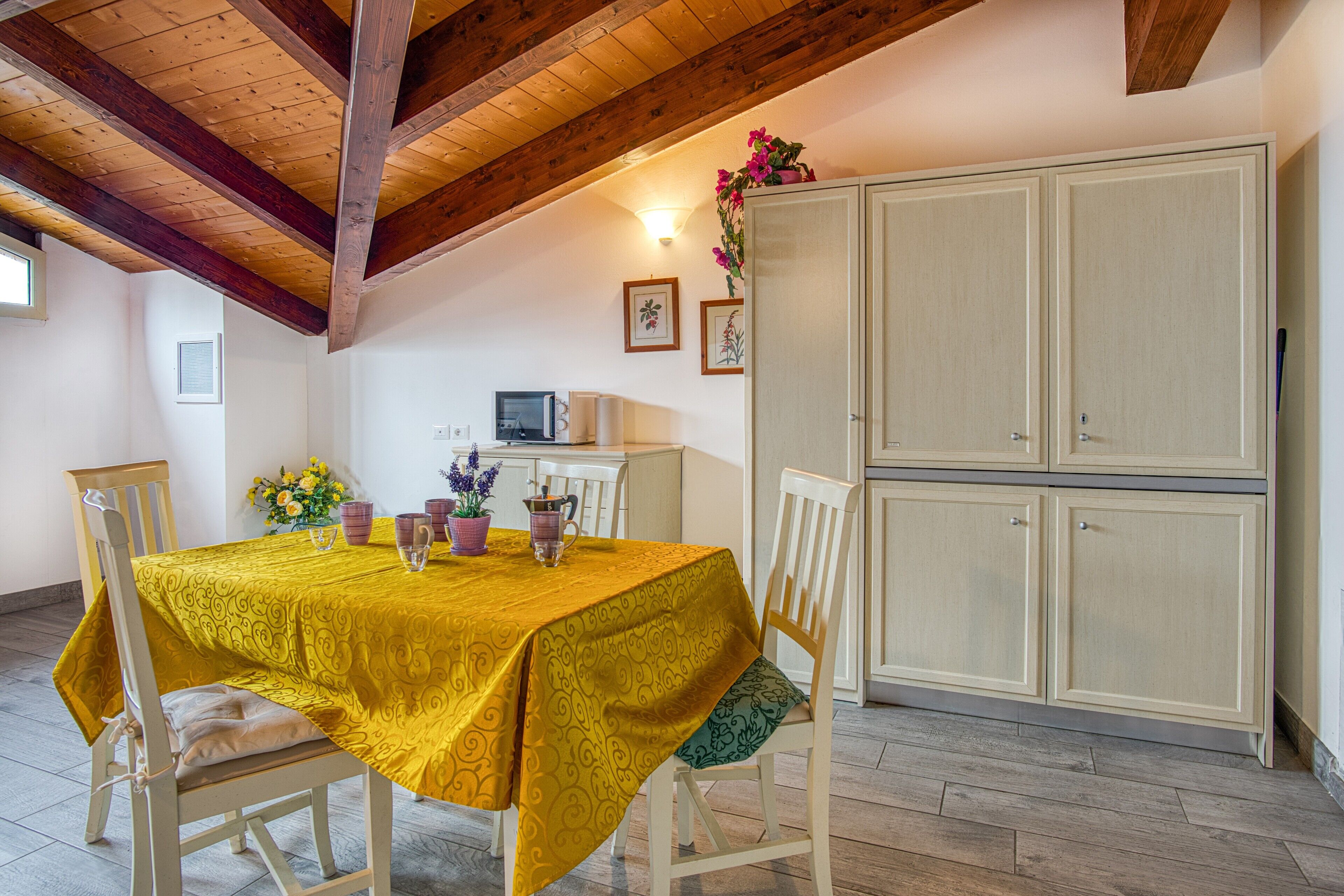 Un coin repas avec une table recouverte d’une nappe jaune, des chaises en bois, un buffet et un four à micro-ondes fixé au mur.