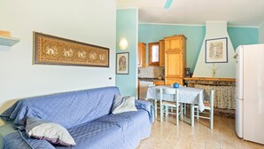 A living room with a blue sofa, a dining table with chairs, and a kitchen area with wooden cabinets and a white refrigerator. - Holiday Home 'Casa Erbì' with Sea View, Private Terrace and Private Garden (Torre dei Corsari Arbus)