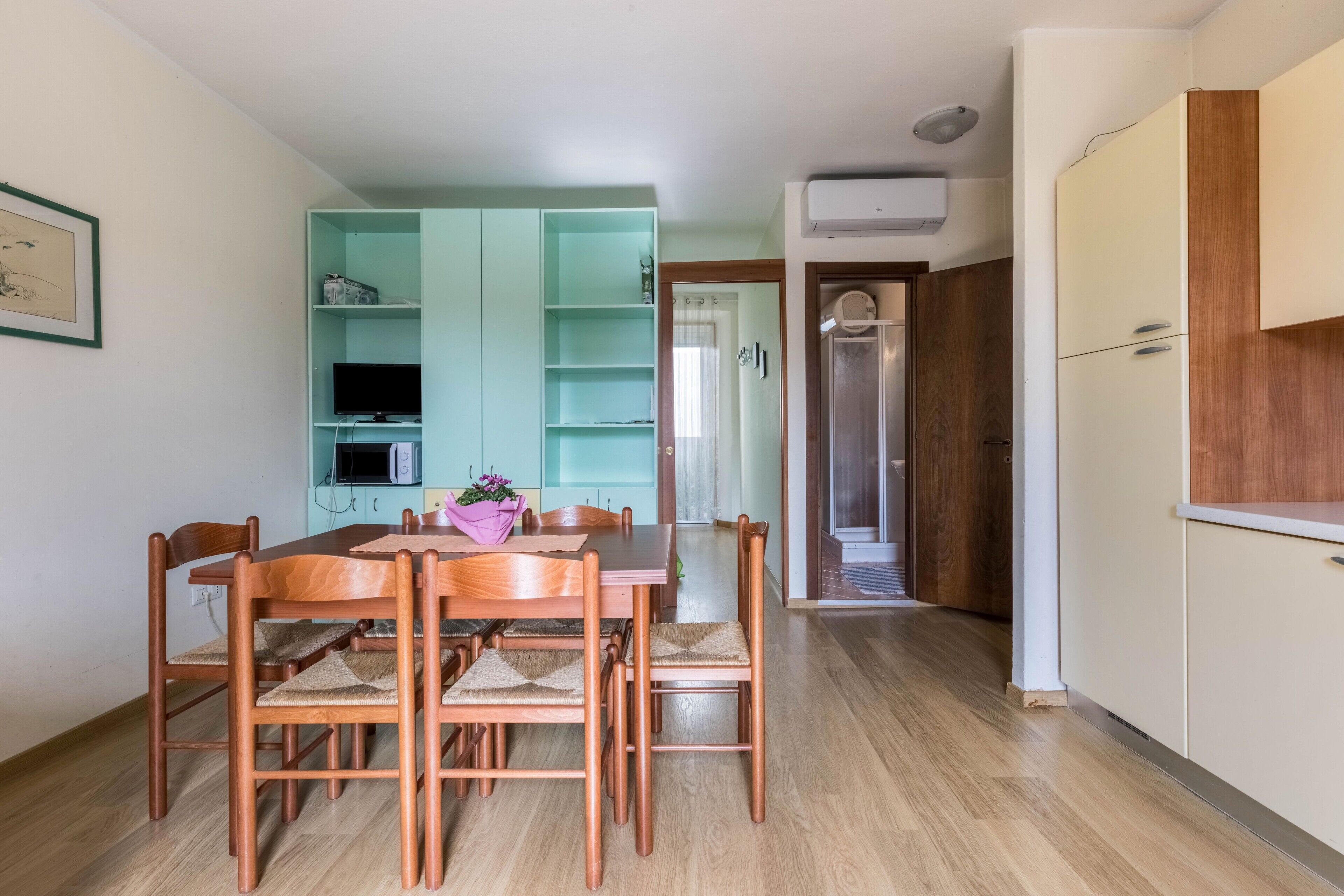 Un comedor con una mesa y sillas de madera, un armario azul verdoso con estantes, una zona de cocina con microondas y una puerta que da a otra habitación.