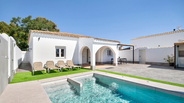 Une piscine à l’eau d’un bleu cristallin, un bâtiment blanc aux portes en arc de cercle et un petit patio aménagé avec des chaises.