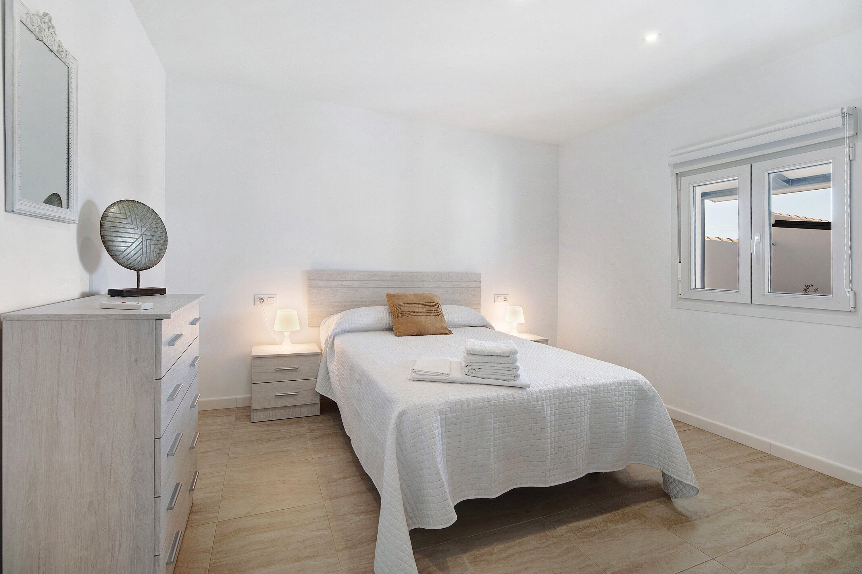 Een slaapkamer met een bed, bedlampen, een dressoir, een spiegel en een raam.