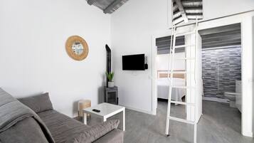 Ein modernes Zimmer mit grauem Sofa, einem kleinen weißen Tisch, einem an der Wand montierten Fernseher, einer Hockerleiter und einem Badezimmer mit gefliestem Duschbereich.