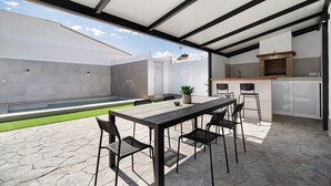 Eine überdachte Terrasse mit Esstisch, Stühlen, einer kleinen Küchenzeile und einem Schwimmbad.