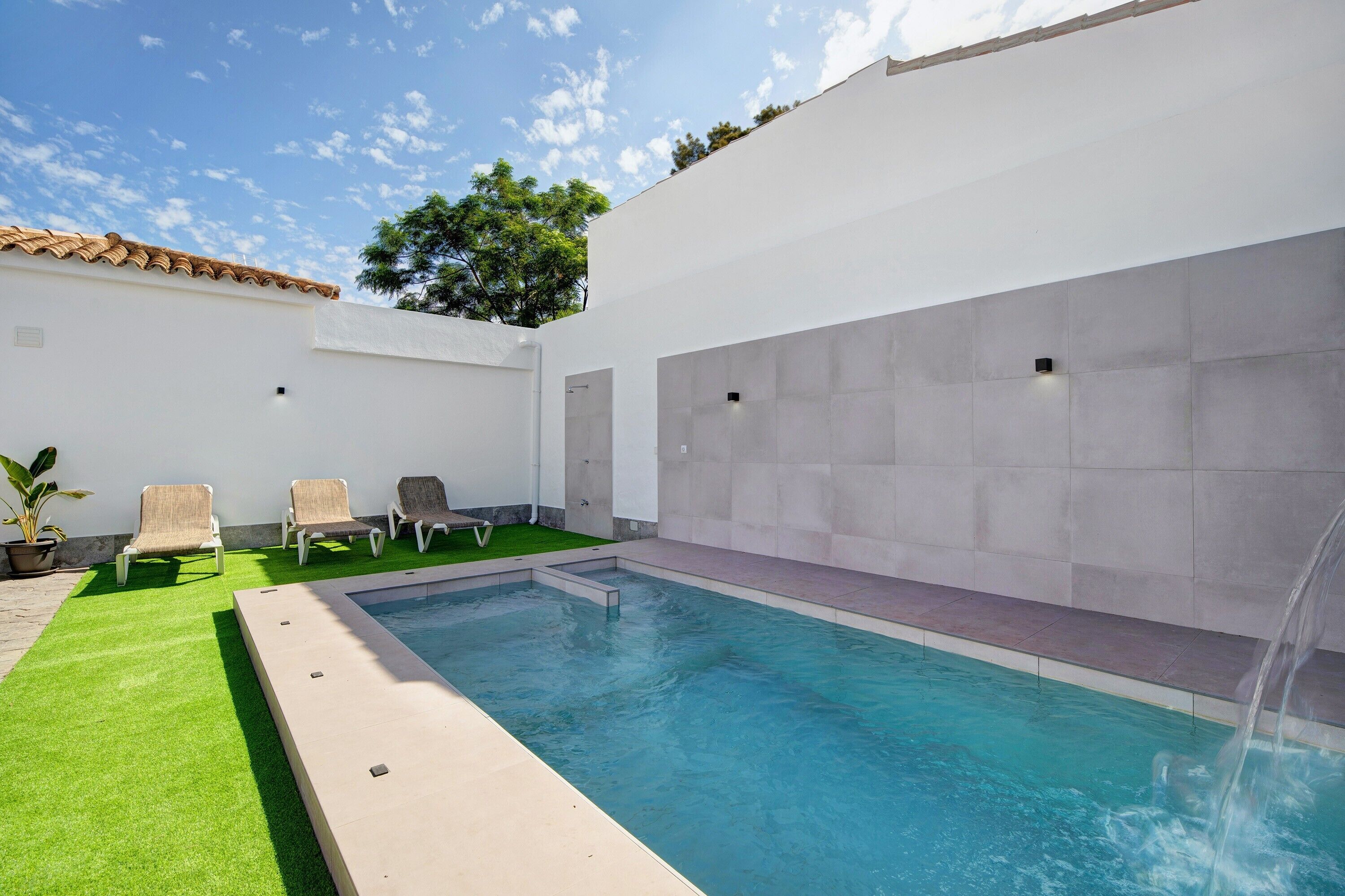 A swimming pool with a tiled deck, a small waterfall feature, and three lounge chairs.