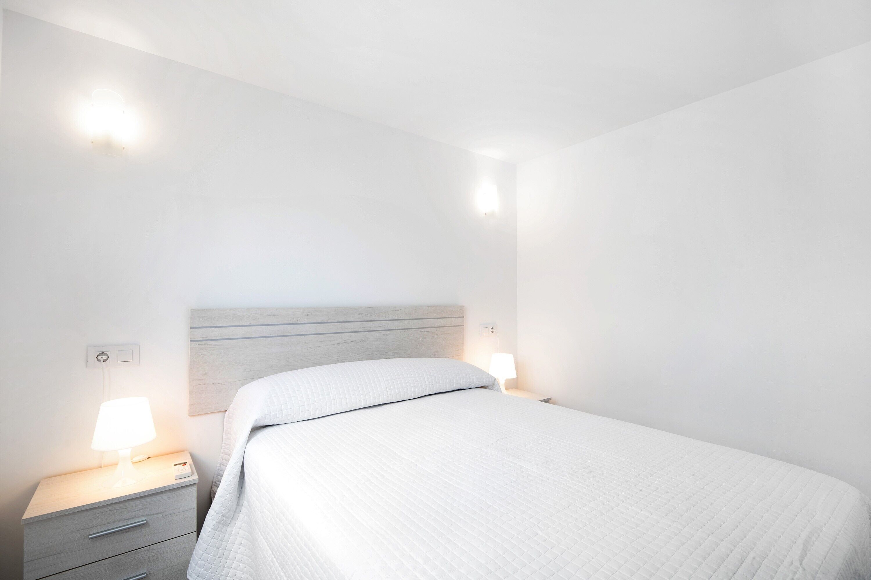 Un dormitorio con cama, lámparas de noche, mesita de noche e iluminación en la pared.